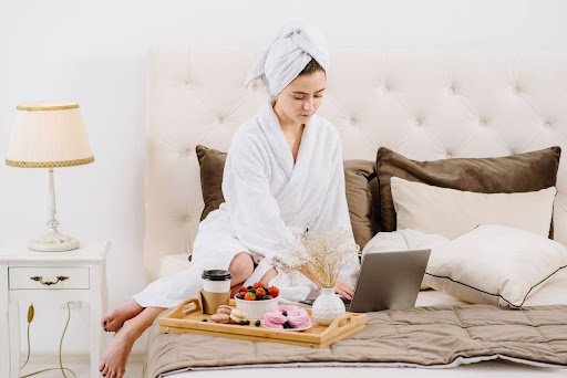 female in bathrobe on bed working on laptop with breakfast tray
